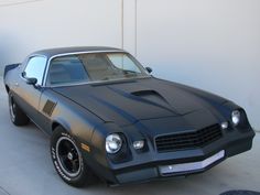 a black muscle car parked in front of a white wall with its hood up and lights on