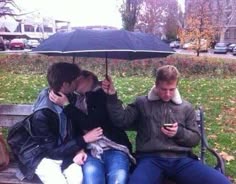 three people sitting on a park bench with an umbrella over their heads and one person holding a cell phone