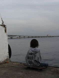 a person sitting on the ground with a skateboard in front of water and bridge