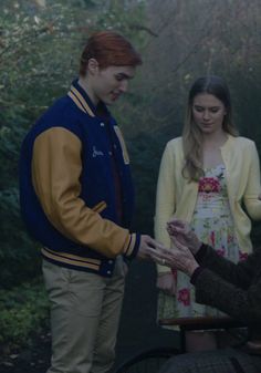 two people standing next to each other in front of trees and bushes, one person is handing something to the other