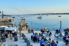 many people are sitting at tables near the water and boats in the water behind them
