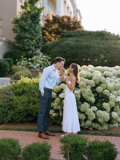 a man and woman standing next to each other in front of some bushes with white flowers