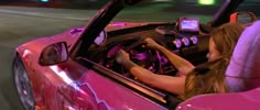 a woman driving a pink sports car down a street at night with her cell phone in the passenger seat