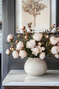 a white vase filled with lots of flowers on top of a marble topped table next to a painting