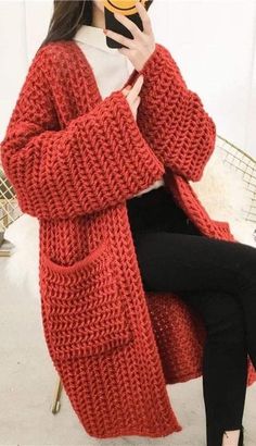 a woman sitting in a chair while holding a cell phone to her ear and wearing a red knitted cardigan