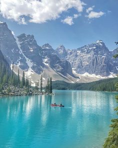 a person in a boat on a lake surrounded by trees and mountains with snow capped peaks