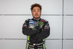 a man with his arms crossed standing in front of a wall wearing a racing uniform