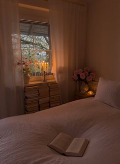 a book is laying on the bed in front of a window with candles and flowers
