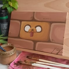 a wooden box with some paint and brushes in it next to a potted plant