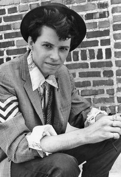 a young man in a suit and hat sitting on the ground next to a brick wall
