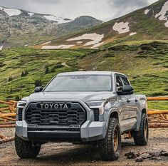 a silver truck parked on top of a dirt road next to green grass covered mountains