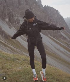 a woman standing on top of a grass covered hillside next to a mountain range with her arms outstretched