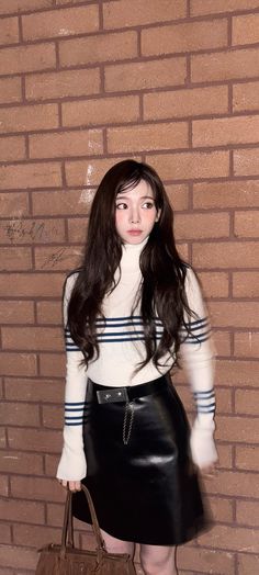 a young woman holding a brown purse standing in front of a brick wall wearing a white sweater and black leather skirt