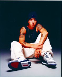 a man sitting on the ground with his arms crossed, wearing sneakers and a hat
