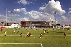 several people playing soccer on a field with clouds in the sky above them and an arena behind them