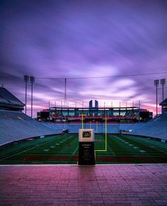 an empty football stadium at dusk with the lights on and purple clouds in the background