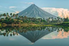 a mountain with trees and clouds reflected in the water near a body of water that is surrounded by palm trees