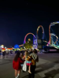some people are walking around at night in front of roller coasters and amusement park rides