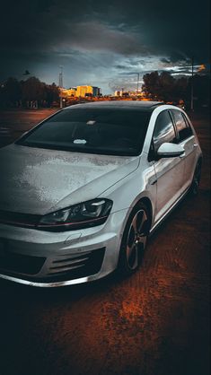 a white car parked in a parking lot with dark clouds above and buildings behind it