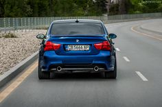 the back end of a blue bmw car driving on a road with trees in the background