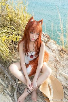 a woman with long red hair sitting on the ground next to some water and wearing a hat