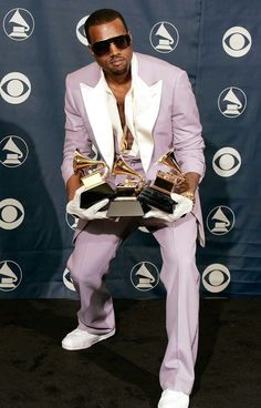 a man in a purple suit and white shirt holding two pairs of shoes while standing on a black carpet