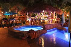 an outdoor swimming pool at night with lights on and tiki bar in the background