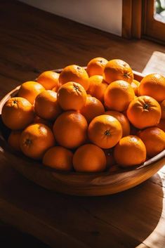a wooden bowl filled with lots of oranges on top of a wood table next to a window