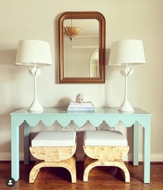 two white lamps sitting on top of a wooden table next to a blue vanity with stools