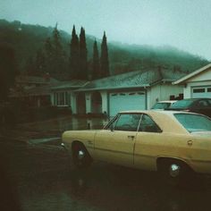 an old yellow car parked in front of a house on a rainy day with no one around