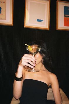 a woman in a black dress holding a martini glass with an olive on it and looking at the camera