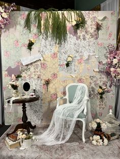 a white chair sitting in front of a wall covered in flowers and greenery next to a table with a birdcage on it
