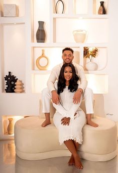 a man and woman sitting on top of a couch in front of shelves filled with vases