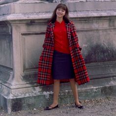 a woman standing in front of a fountain wearing a red shirt and plaid coat with black heels