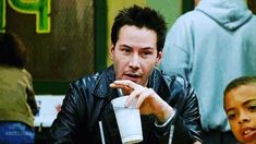 a man in a leather jacket holding a cup and pointing to the side while sitting at a table