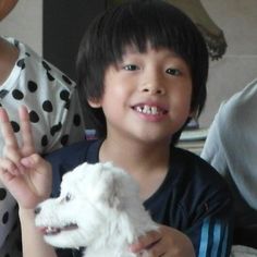 a young boy holding a white dog in his hand and smiling at the camera with two other people behind him