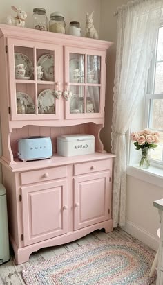 a pink china cabinet with glass doors and drawers in a room next to a window