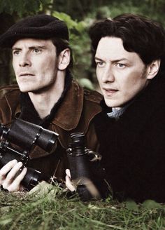 two men sitting in the grass looking at something with binoculars on their hands and one holding a camera