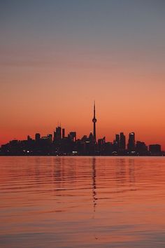 the city skyline is reflected in the calm water at sunset, with an orange glow on the horizon
