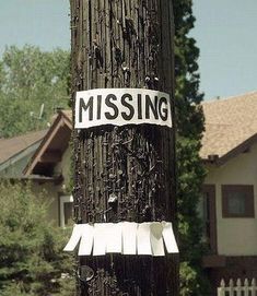 a missing sign on a telephone pole in front of a house with trees and houses behind it