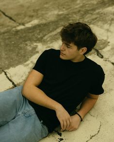 a young man sitting on the ground with his hands in his pockets and looking off into the distance