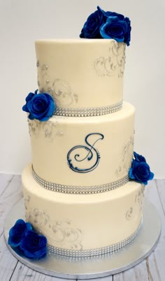 a three tiered wedding cake with blue roses on the side and monogramming