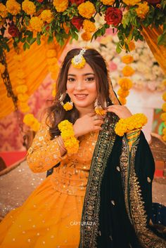 a woman in yellow and black dress sitting under a flower covered arch with flowers on it