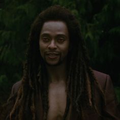 a shirtless man with dreadlocks standing in front of some trees and looking at the camera