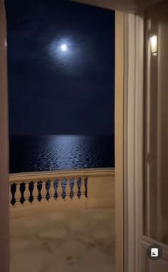 an open door leading to a balcony overlooking the ocean at night with a full moon in the sky