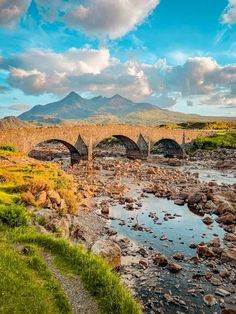 an old stone bridge over a river with mountains in the backgrouund and clouds in the sky