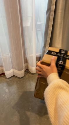 a person holding a brown box in front of a window with curtains behind it and a white curtain