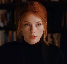 a woman with red hair and blue eyes stares into the camera while standing in front of bookshelves