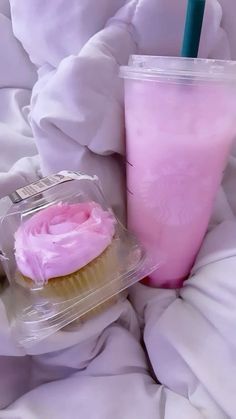 a cupcake with pink frosting next to a drink in a plastic container on a bed