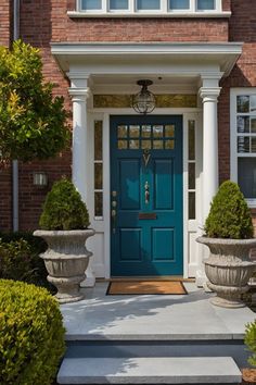 A charming entryway featuring a teal front door, flanked by two ornate planters with shrubbery, set against a brick facade.